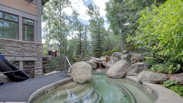 Un jacuzzi rodeado de rocas grandes y vegetación, con un muro de piedra y una barandilla de metal.