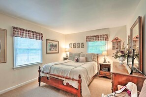 Quarto com cama de madeira, duas janelas com cortinas xadrez, mesa de cabeceira com abajur, cômoda e espelho.