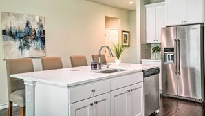Cocina moderna con armarios blancos, un refrigerador de acero inoxidable y un comedor con una isla blanca y sillas.