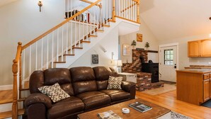A living room with a brown leather sofa, wooden staircase, and a brick fireplace. - Waterfront Sebec Lake Home w/ Yard + Fire Pit (Dover-Foxcroft)