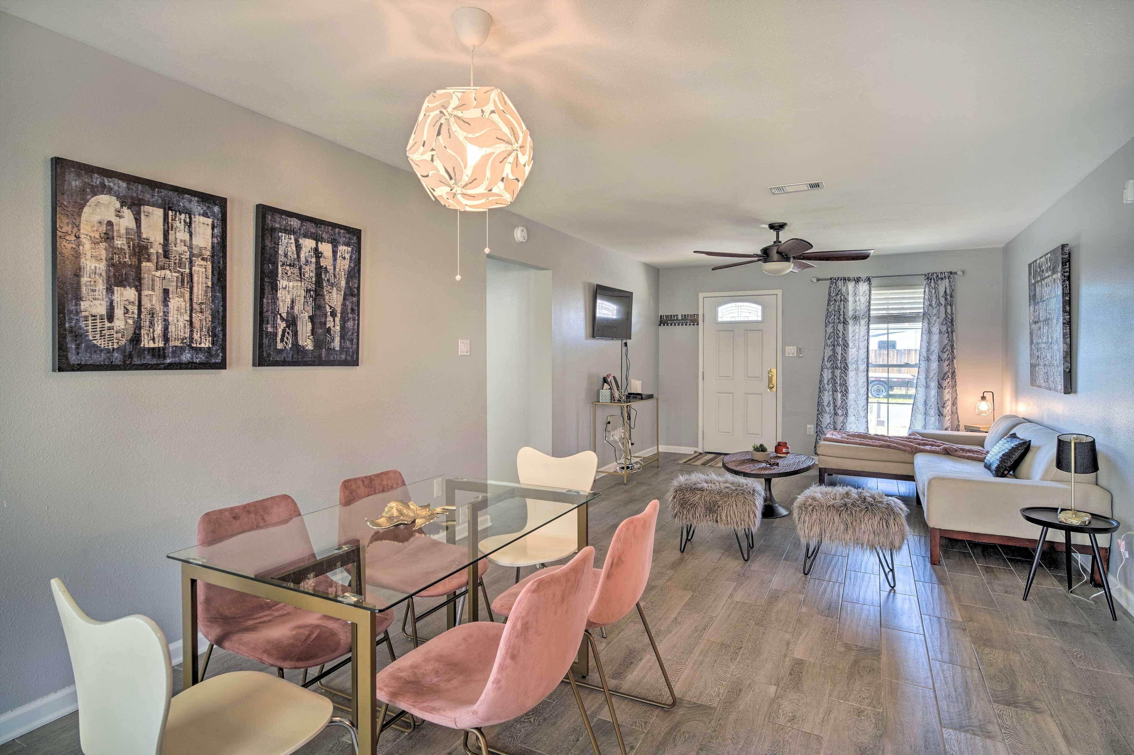 A modern living room with a glass dining table, pink chairs, a sofa, and a ceiling fan.