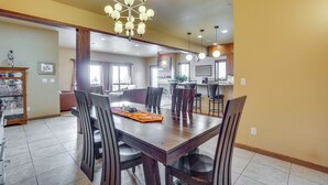 A spacious dining area with a large wooden table and chairs, a chandelier, and a kitchen in the background. - Upscale Laramie Home w/ Hot Tub & Patio! (Laramie)