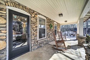A stone exterior with a black door, a covered porch with a rocking chair, and a view of a snowy landscape. - Quaint Home w/ Porch in Downtown Waynesville! (Waynesville)