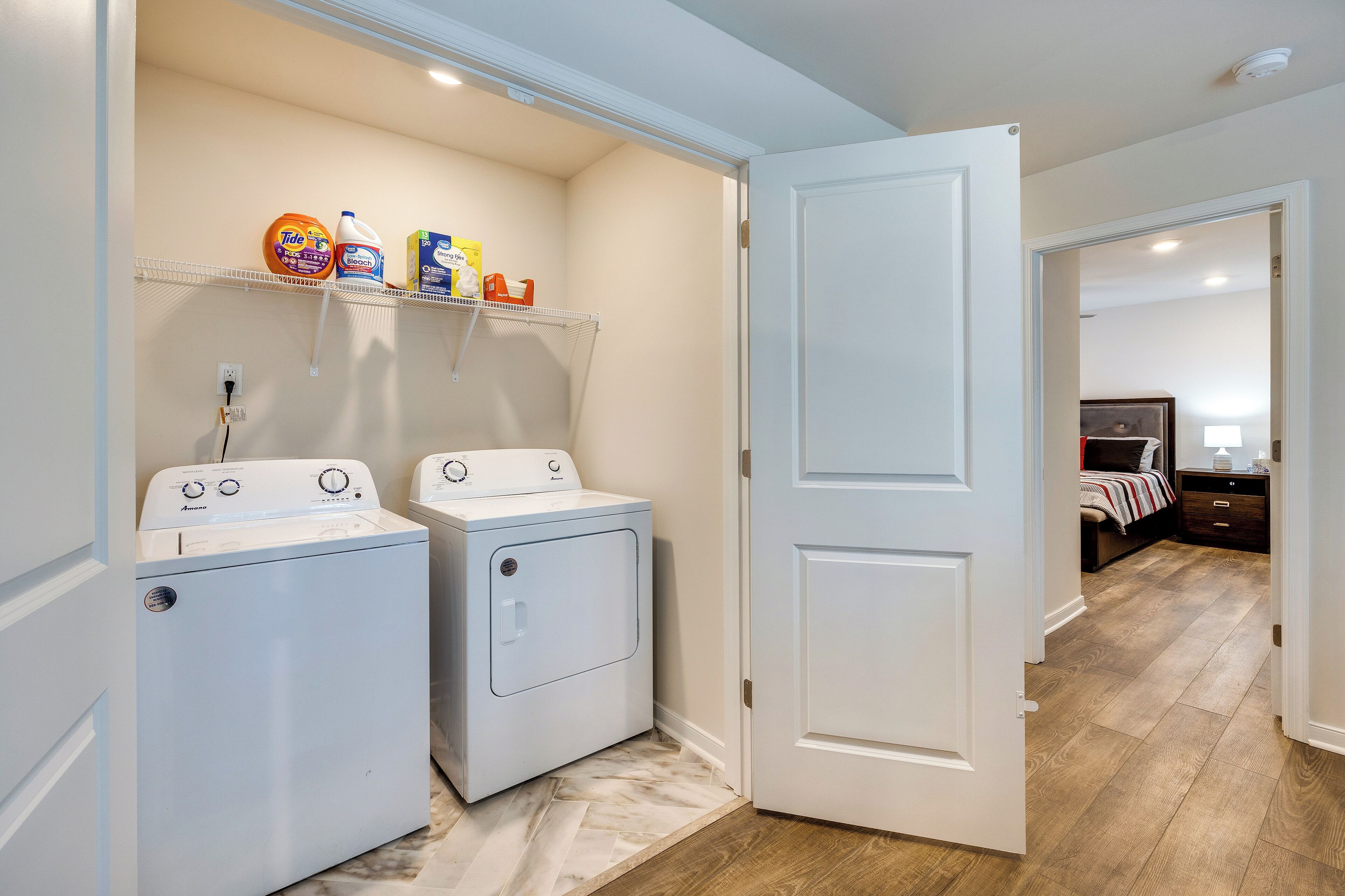 A laundry area with two white front-loading washing machines, a shelf with cleaning supplies, and a door leading to a bedroom.