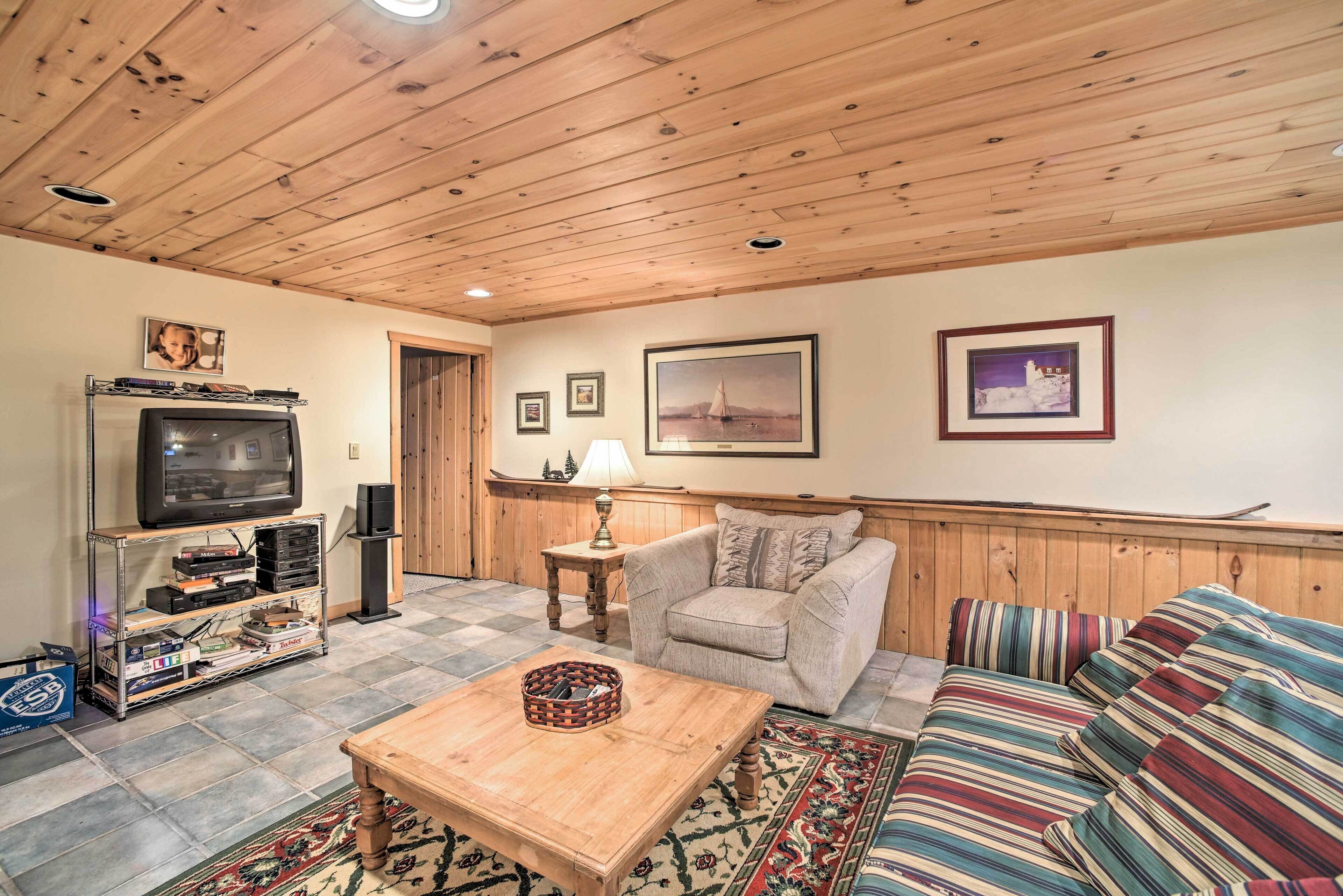 A cozy living room with a wooden ceiling, a television on a shelf, a sofa, a patterned rug, and framed pictures on the wall.