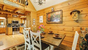 A wooden interior with a dining table set for four, surrounded by white chairs. A kitchen with wooden cabinets and a microwave is visible in the background.
