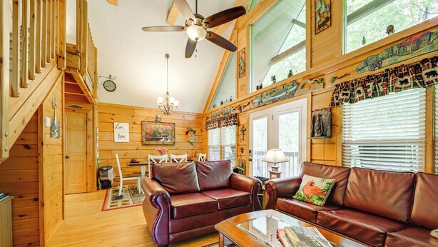 A cozy cabin interior with a wooden staircase, a brown leather sofa, a glass coffee table, and a ceiling fan.