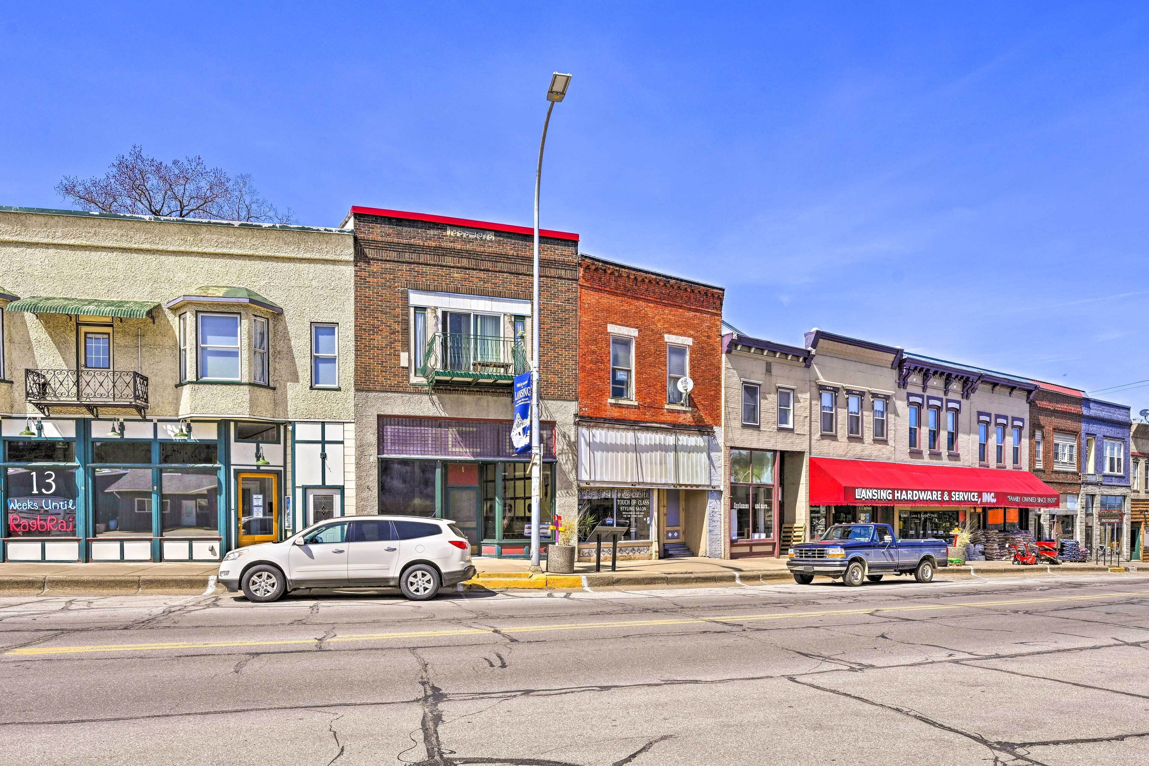 A street view with a white SUV parked on the side of the road, a red building with a sign for 'LANSING HARDWARE & SERVICE, INC.', and a storefront with a sign that reads '13 Weeks Until Raspbari'.