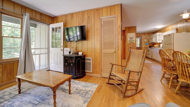 Una habitación acogedora con piso de madera, un televisor montado en la pared, una mecedora, una mesa de comedor y una zona de cocina con armarios y electrodomésticos.