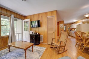 A cozy room with a wooden floor, a television mounted on the wall, a rocking chair, a dining table, and a kitchen area with cabinets and appliances.