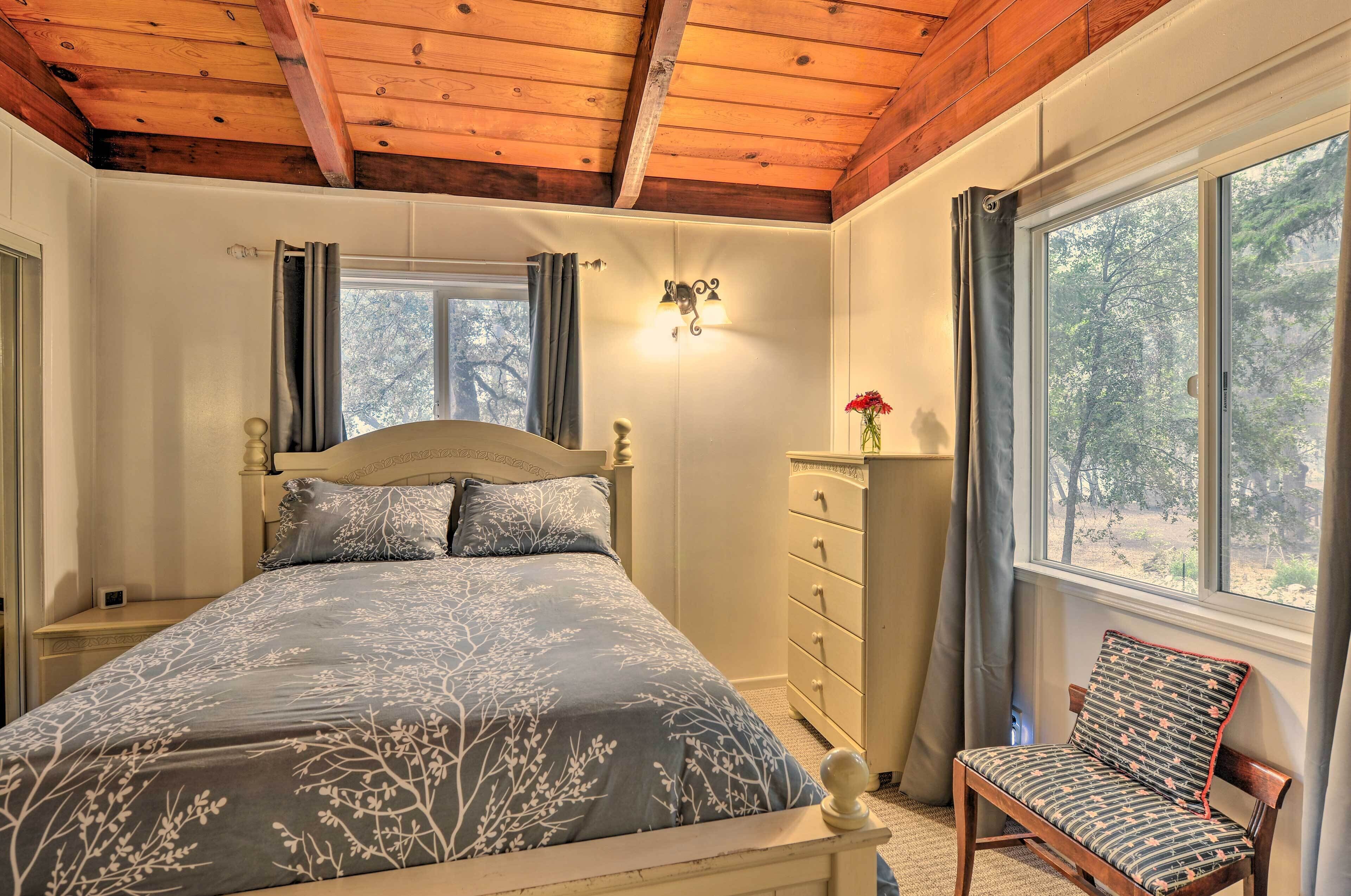 Un dormitorio con techo de madera, una cama con colcha estampada, una mesita de noche, un aparador, una silla y una ventana con vistas a árboles.