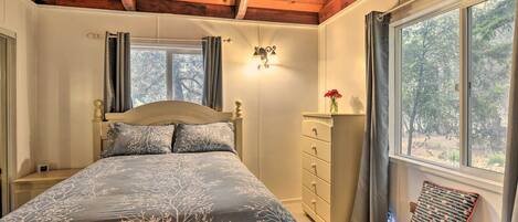 Un dormitorio con techo de madera, una cama con colcha estampada, una mesita de noche, un aparador, una silla y una ventana con vistas a árboles.