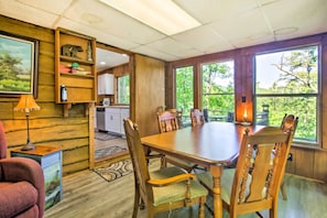 Un comedor con mesa y sillas de madera, una cocina al fondo y grandes ventanales con vistas a zonas verdes.