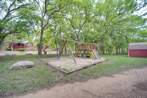 A playground with swings and slides, surrounded by trees and a red building in the background. - Winery Cabin in Fredericksburg w/ Wraparound Deck! (Fredericksburg)