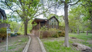 A rustic cabin with a wooden deck and a covered porch, surrounded by trees and a gravel path. - Winery Cabin in Fredericksburg w/ Wraparound Deck! (Fredericksburg)