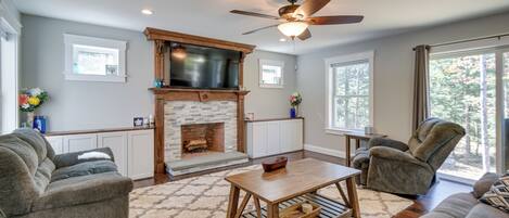 A living room with a fireplace, a ceiling fan, and a large window.