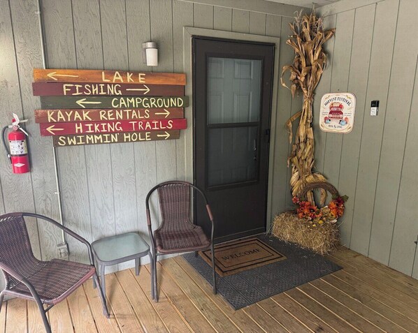 A wooden porch with a welcome mat, a fire extinguisher, and a signboard with directions to various amenities like a lake, fishing, and a swimming hole.
