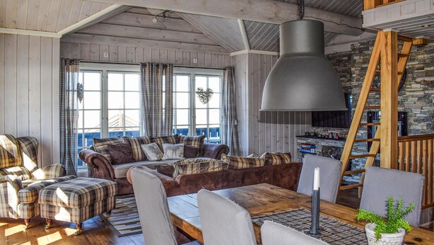Ein gemütliches Wohnzimmer mit einer Couch, Sesseln, einem Esstisch und einem großen Fenster mit Vorhängen.