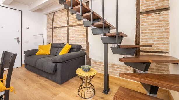 A modern living room with a wooden staircase, a grey sofa with yellow cushions, and a small round table with a bowl. - Castello Loft Heaven (Cagliari)