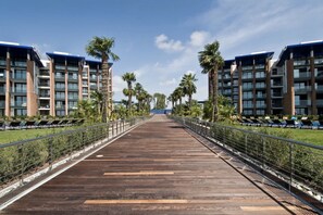 A long, wooden pathway flanked by modern buildings with balconies, leading to a distant view. - Frontemar Residenze sul mare D6 - Carrraro Immobiliare Jesolo (Lido di Jesolo)