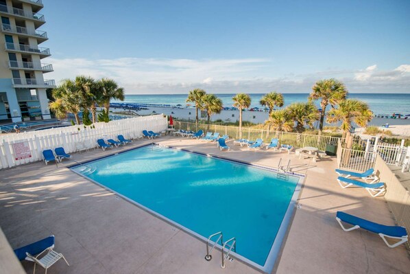 A swimming pool with blue chairs and palm trees, overlooking a beach.