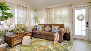 Un salon chaleureux avec un canapé marron, des coussins décoratifs, un panier en osier et un tapis aux motifs tropicaux.