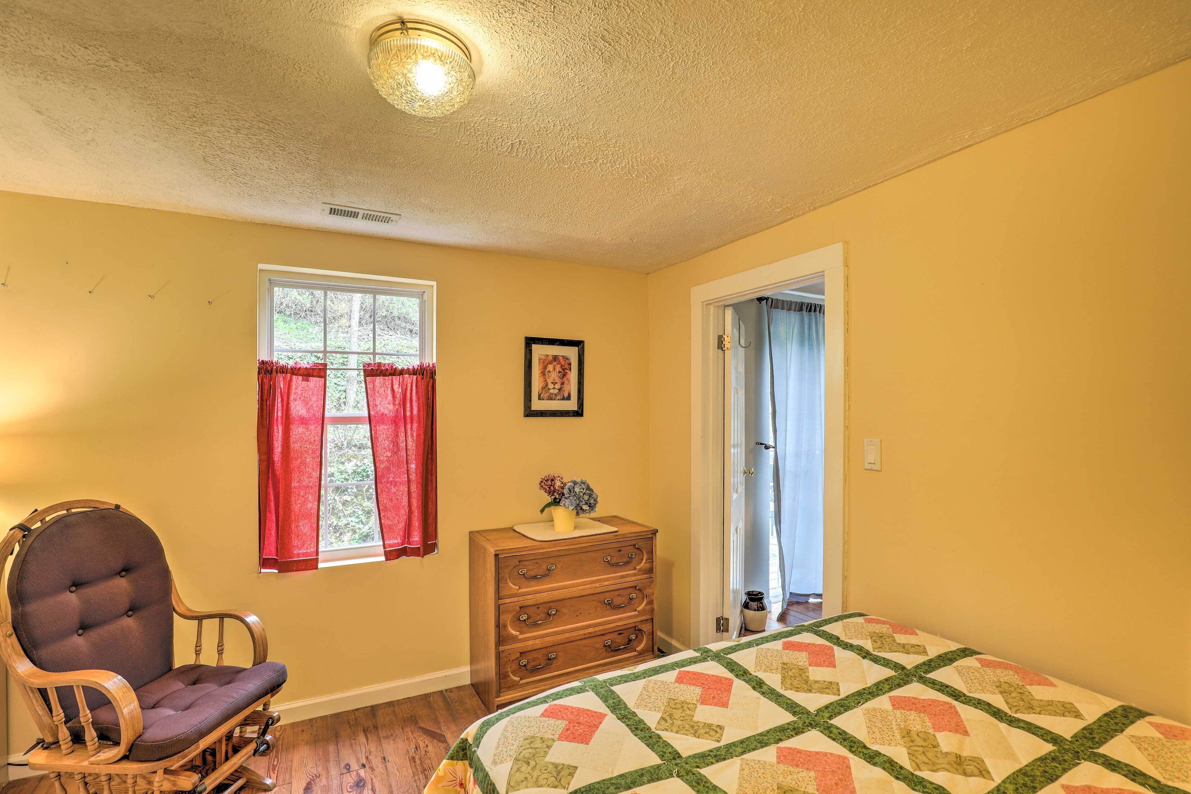 A bedroom with a wooden rocking chair, a dresser, a quilted bedspread, and a window with red curtains.