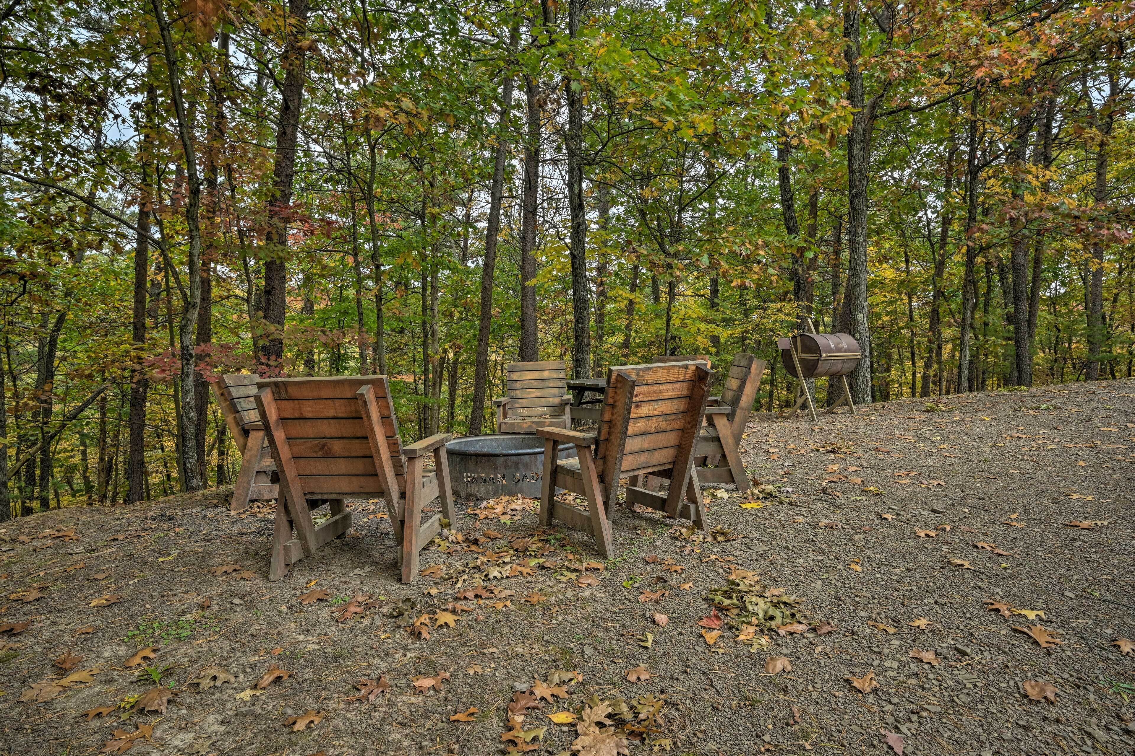 A rustic outdoor seating area with wooden benches and a table, surrounded by a forest with autumn foliage.