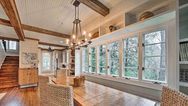 A spacious dining area with a wooden table and wicker chairs, large windows with a view of trees, and a staircase leading to another level. - Beautiful Trillium Home Next to Waterfall! (Cashiers)