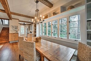 Uma sala de jantar espaçosa com mesa de madeira e cadeiras de fibra, grandes janelas com vista para árvores e uma escada que leva a outro andar.