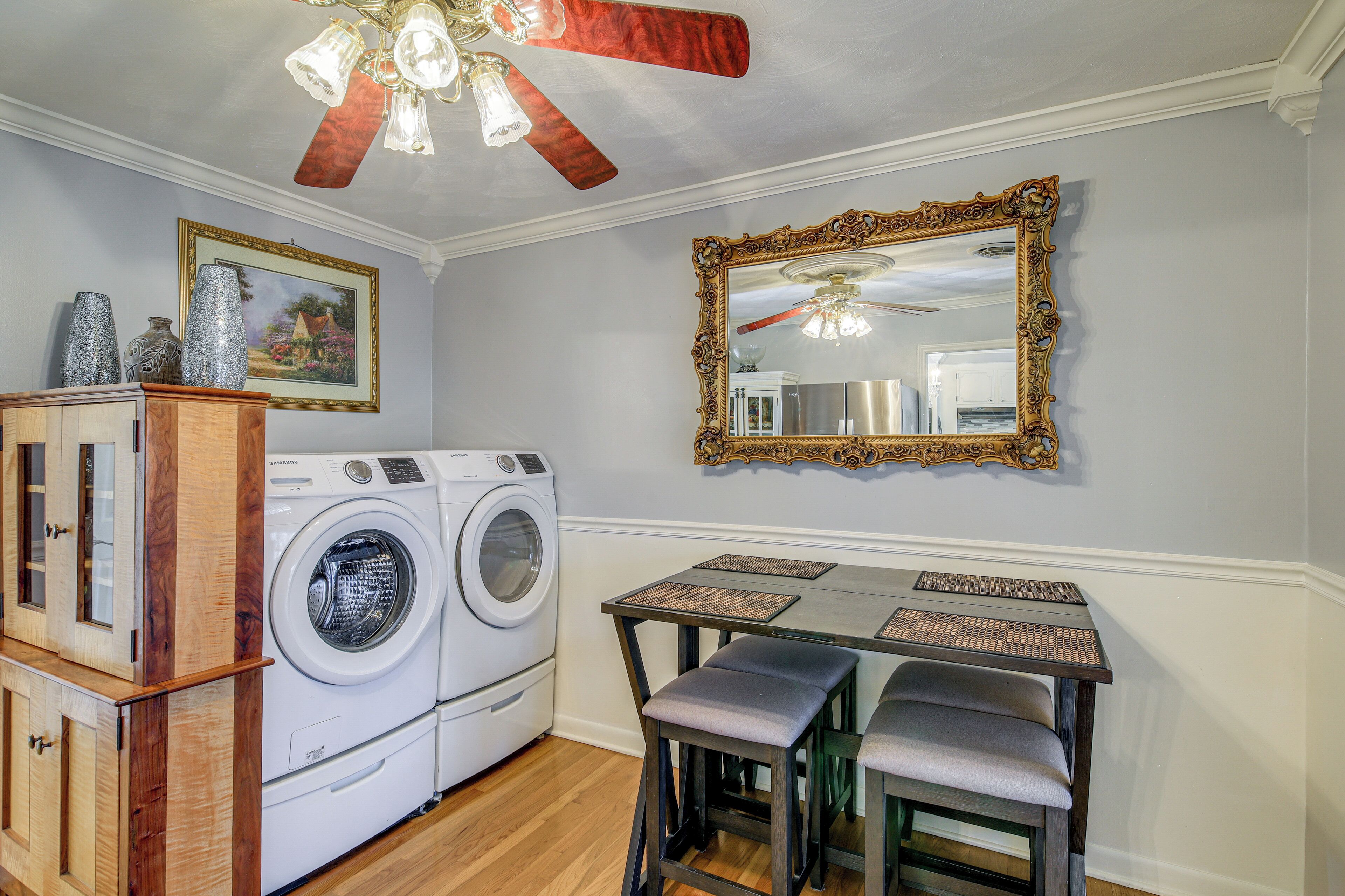A laundry area with a washing machine, a wooden cabinet, a dining table with two chairs, and a large ornate mirror.
