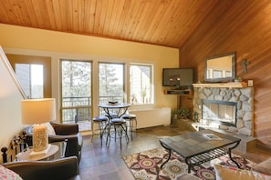 A cozy living room with a stone fireplace, a dining area with a table and chairs, and a television mounted on the wall. - Breathtaking Bend Condo w/ Resort Amenities! (Bend)