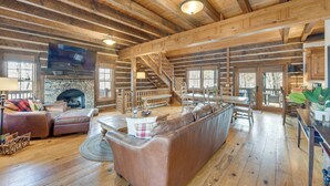 A spacious log cabin interior with a stone fireplace, leather sofas, wooden floors, and a staircase leading to an upper level.
