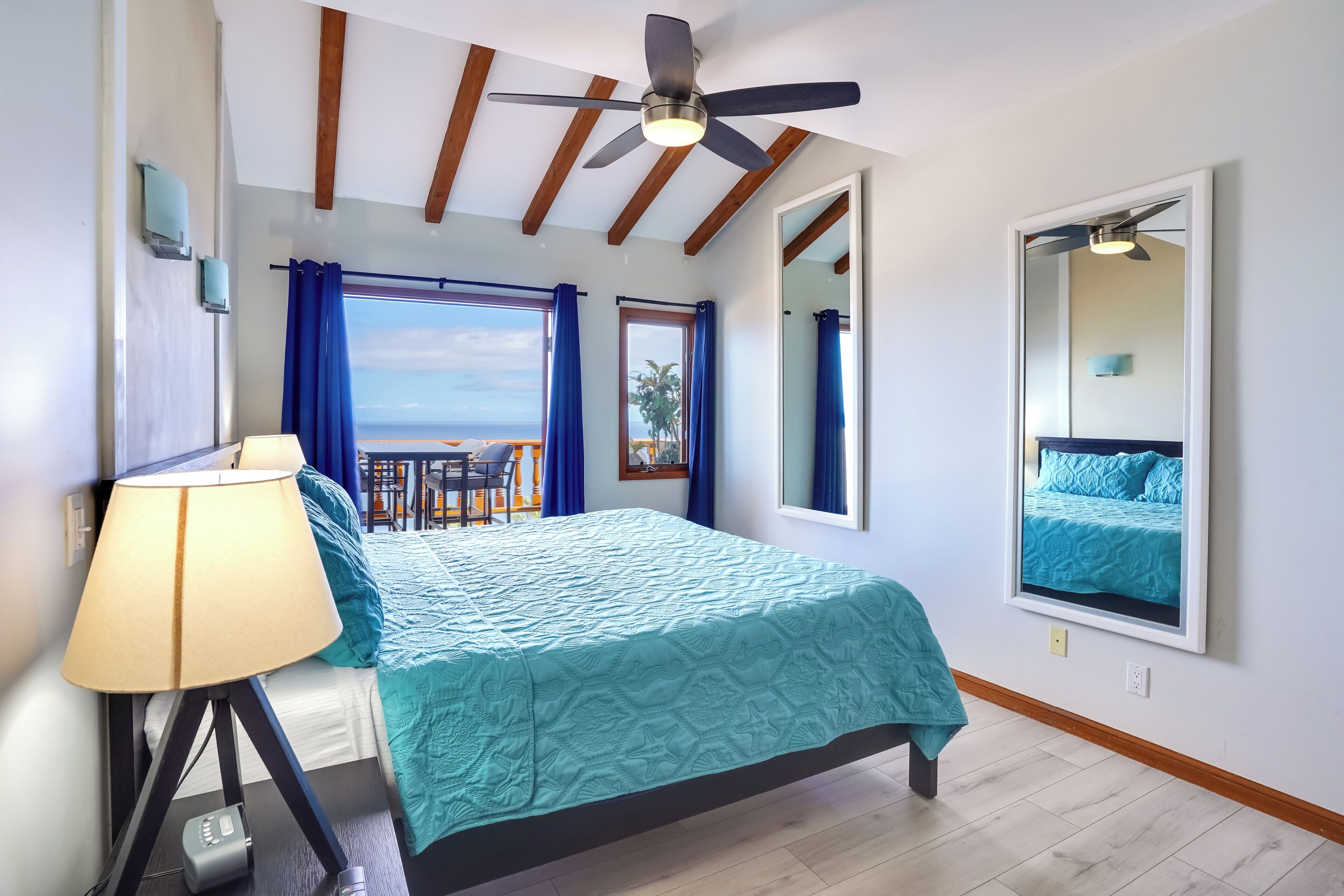 A bedroom with a bed, a ceiling fan, a nightstand, a lamp, and a view of the ocean through large windows.