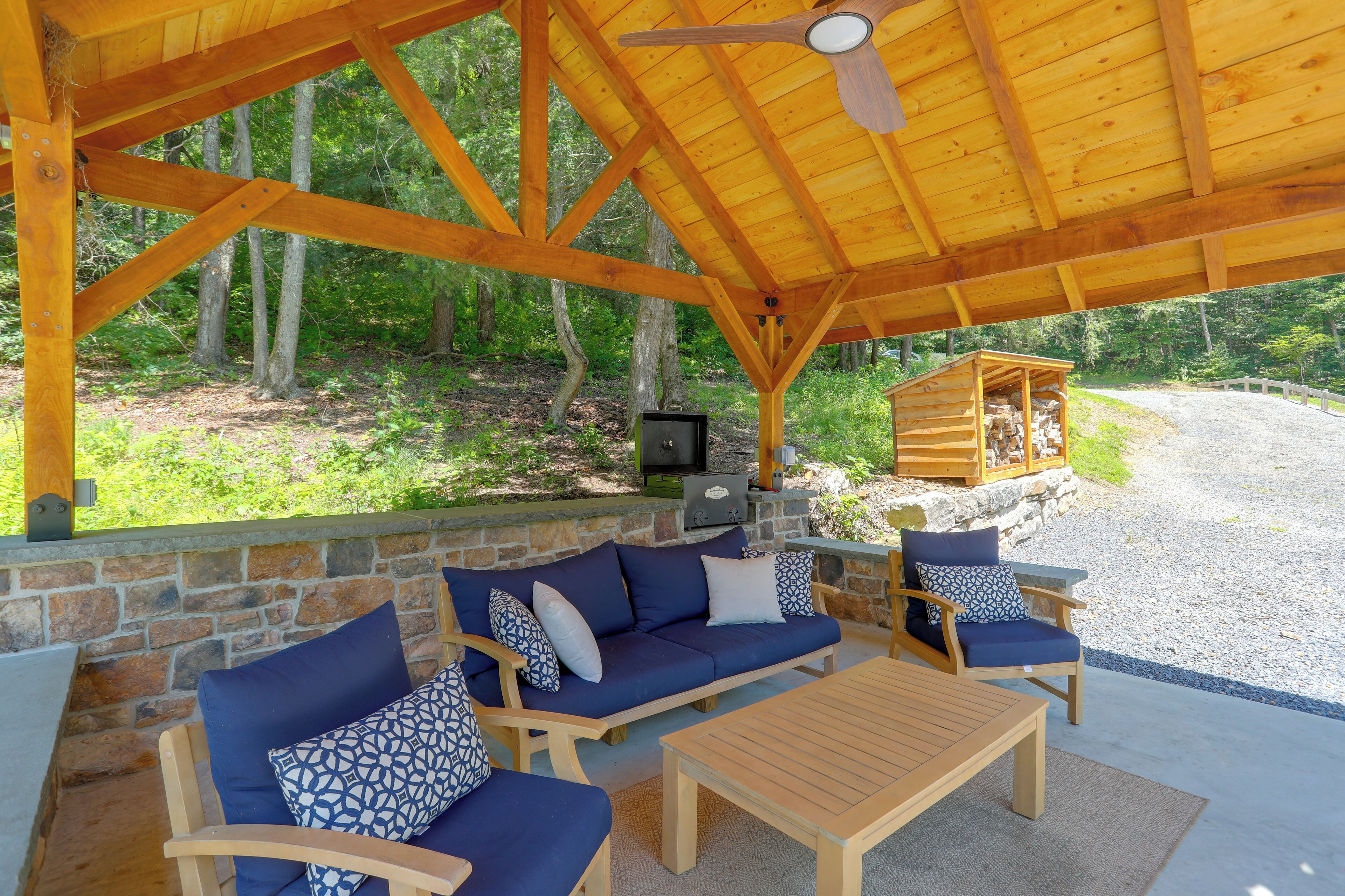 Un espace extérieur aménagé avec des sièges, abrité par un toit, doté d’un mur en pierre, de mobilier en bois et offrant une vue sur la forêt.
