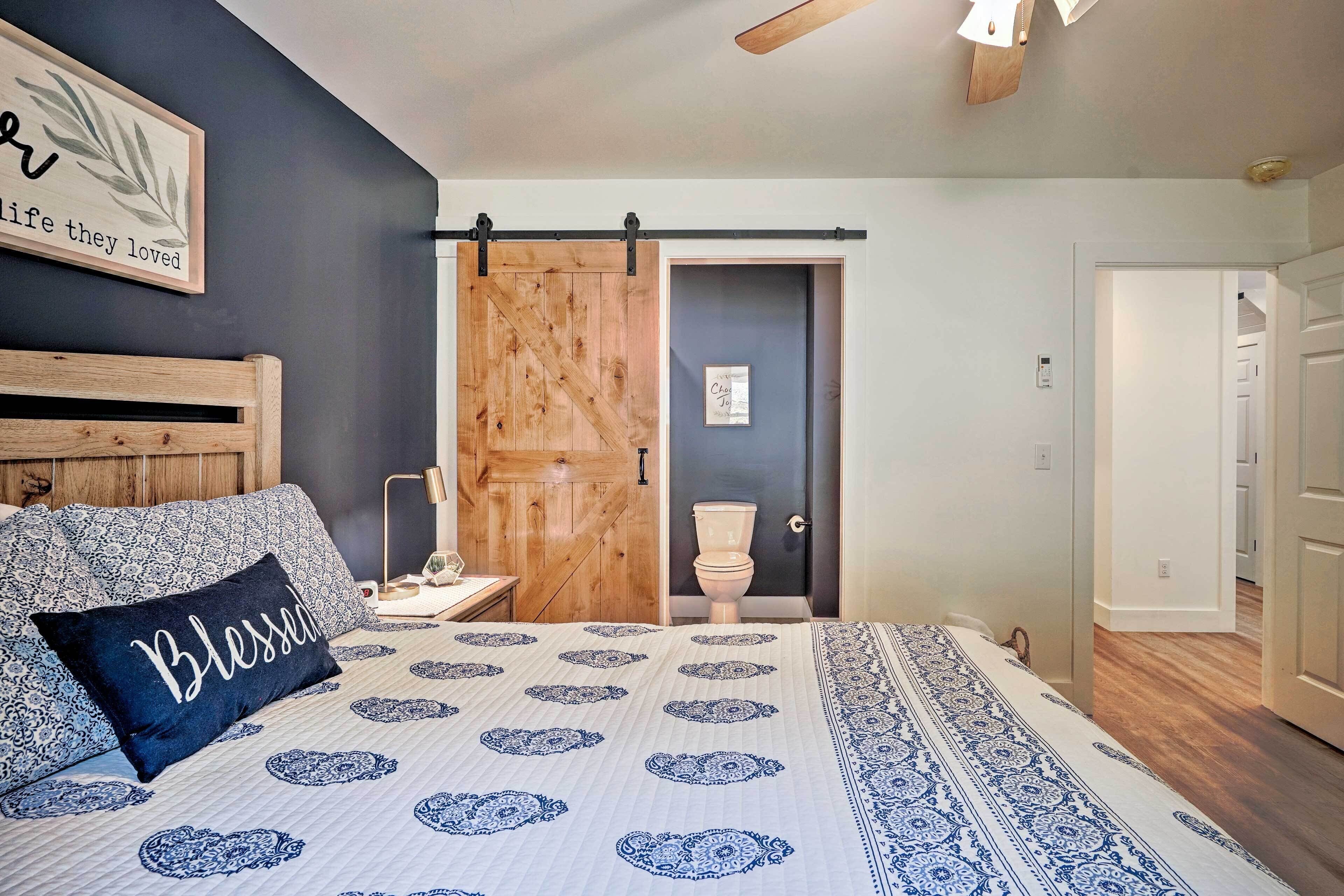 A bedroom with a bed, a nightstand, a wooden door, and a bathroom visible through an open door.