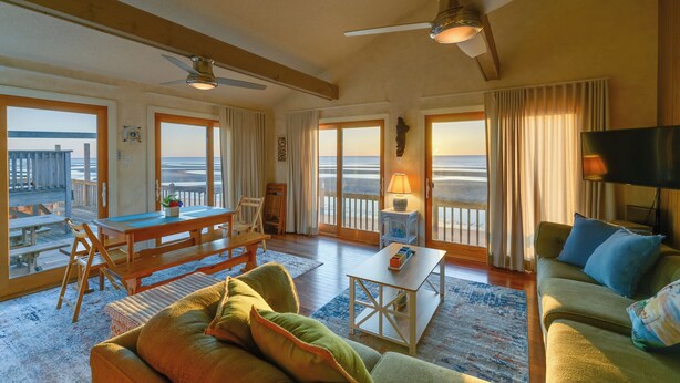 A spacious living room with a large window overlooking the beach, a ceiling fan, and a cozy seating area with a sofa and a table. - Delaware Bay & Sunset Views: Cape May Cottage (Cape May Court House)