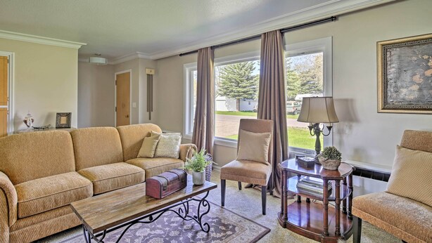 A living room with a beige sofa, a wooden coffee table, and a chair. There is a window with curtains, a lamp on a side table, and a framed picture on the wall. - Ashton Escape w/ Yard: 56 Mi to West Yellowstone! (Ashton)