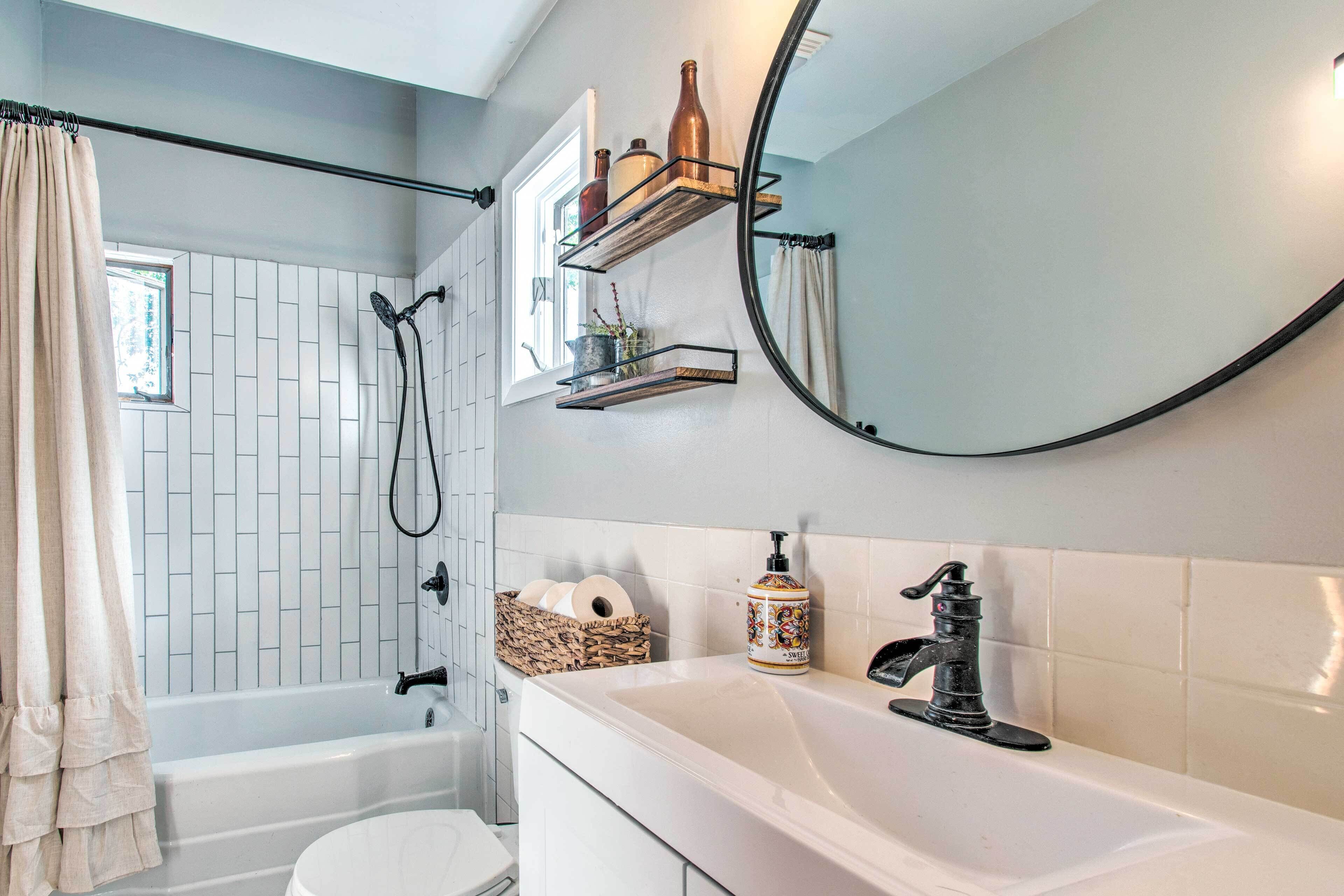 Une salle de bain avec une baignoire, des toilettes et un lavabo. Il y a une fenêtre avec un rideau, un miroir et des étagères avec des bouteilles et un panier.