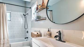 Une salle de bain avec une baignoire, des toilettes et un lavabo. Il y a une fenêtre avec un rideau, un miroir et des étagères avec des bouteilles et un panier.