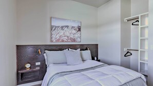 A bedroom with a bed, a nightstand, a wall-mounted shelf, a hanging coat rack, and a painting.