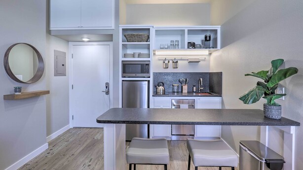 A compact kitchen with white cabinetry, a built-in microwave, and a coffee machine. There are two white chairs and a small table.