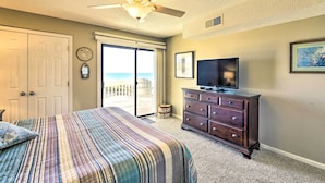 Ein Schlafzimmer mit Bett, Kommode, Fernseher und Blick auf den Strand durch eine Schiebetür.