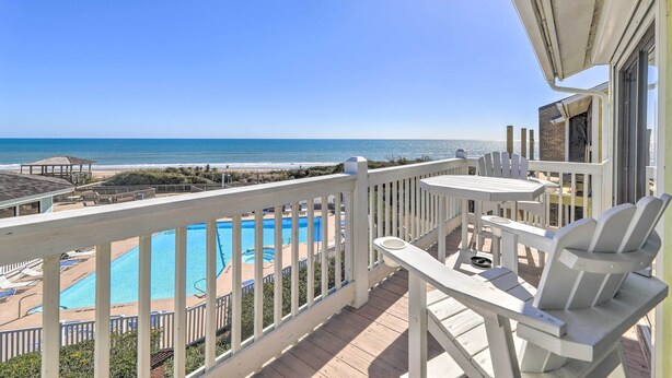 Ein Balkon mit Blick auf ein Schwimmbecken und einen Strand, weiße Gartenmöbel und ein klarer, blauer Himmel.