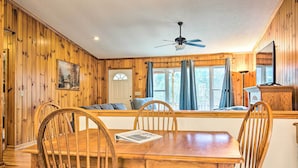 A wooden-paneled room with a dining table, chairs, a ceiling fan, and a window with curtains.