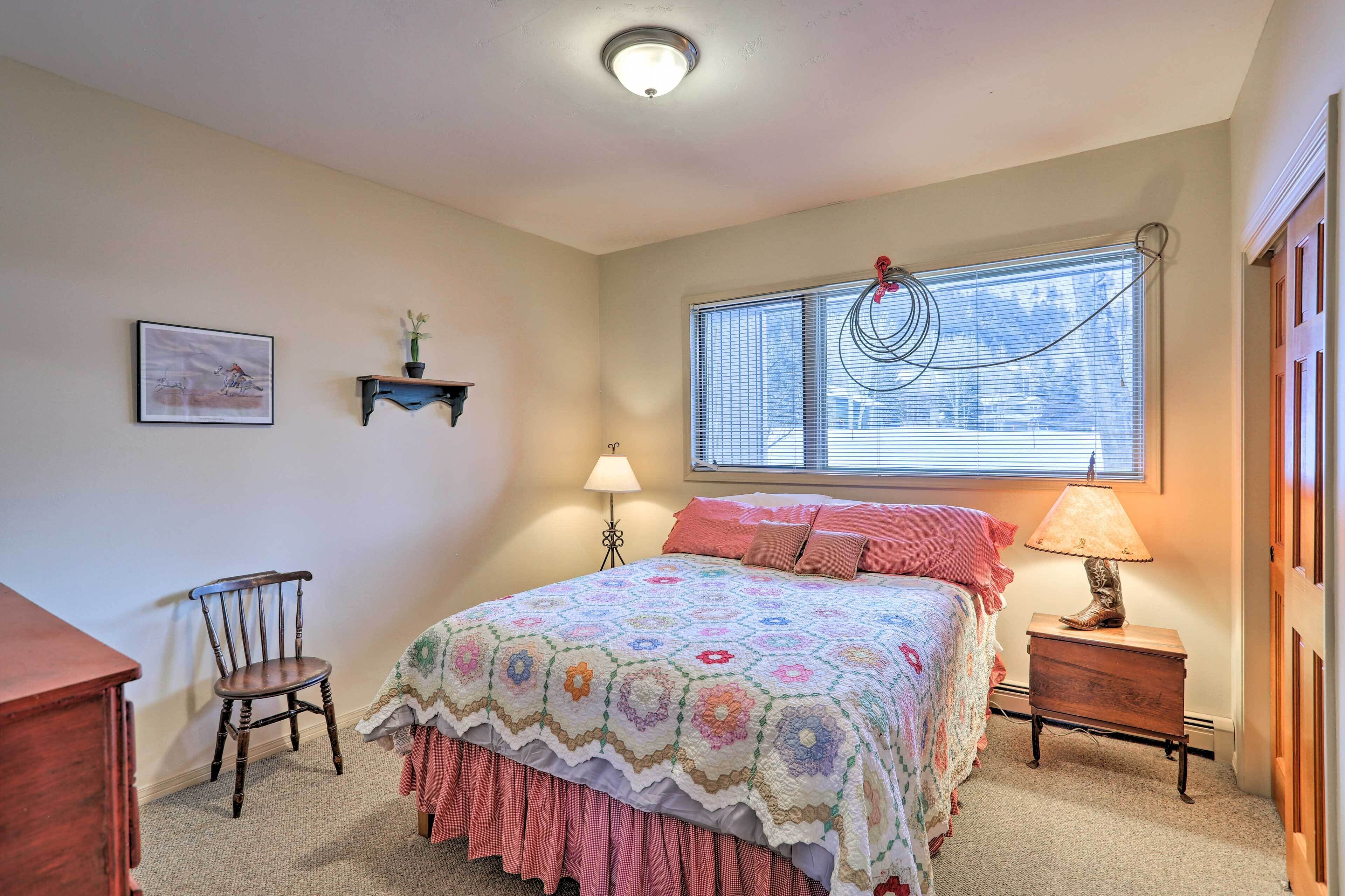A bedroom with a bed, a chair, a nightstand, a lamp, a window with blinds, and a picture on the wall.