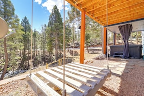 A wooden deck with a view of a forest, a hot tub, and a covered area with a bench. - Colorado Cabin Getaway Near Pikes Peak! (Woodland Park)