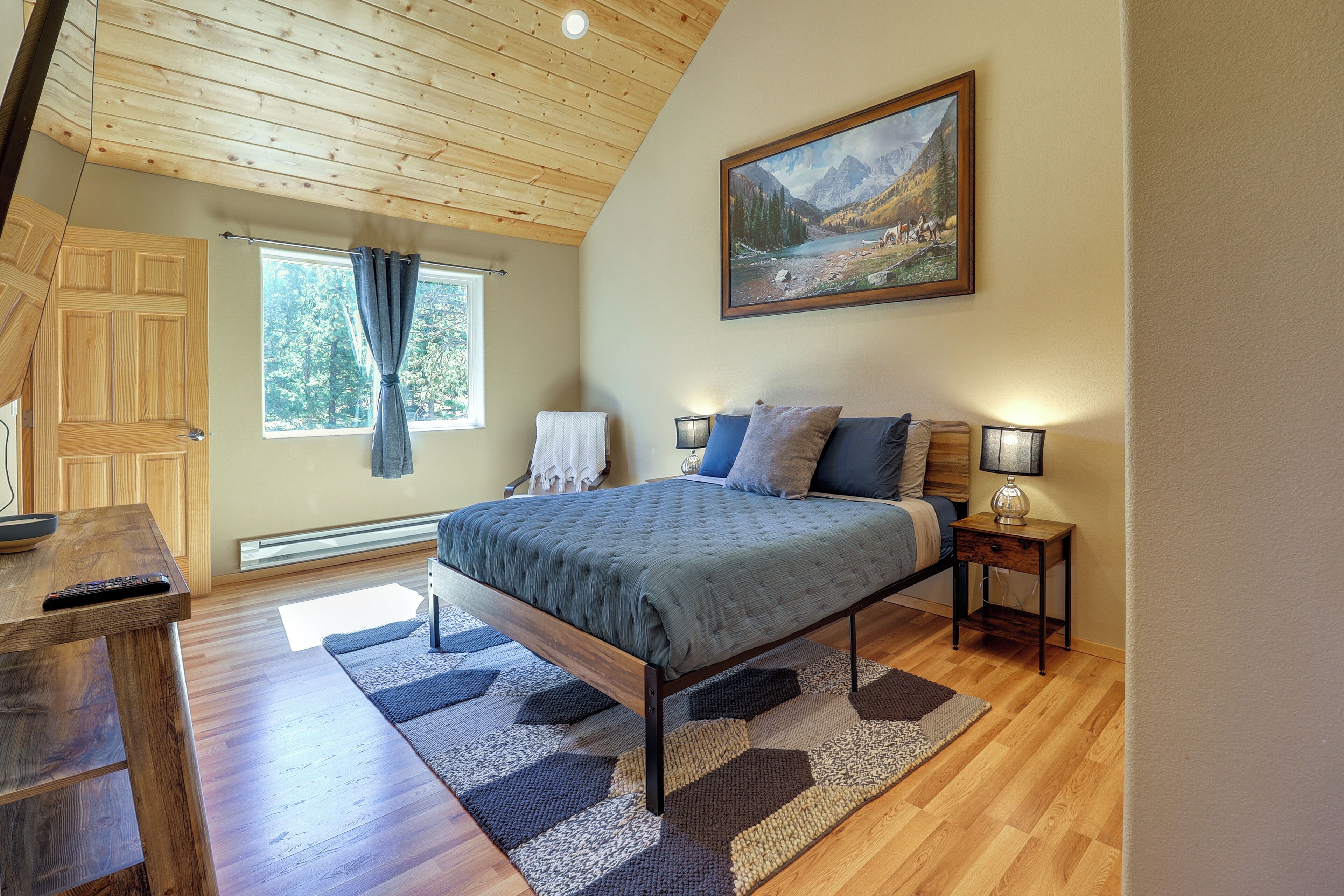Un dormitorio con techo de madera, una cama con manta gris, una mesita de noche con lámpara y un cuadro de un paisaje montañoso.