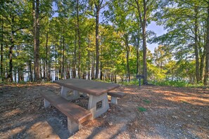 A picnic table in a wooded area with a lake in the background. - Coffeeville Cabin w/ Deck ~ 7 Mi to Grenada Lk (Coffeeville)