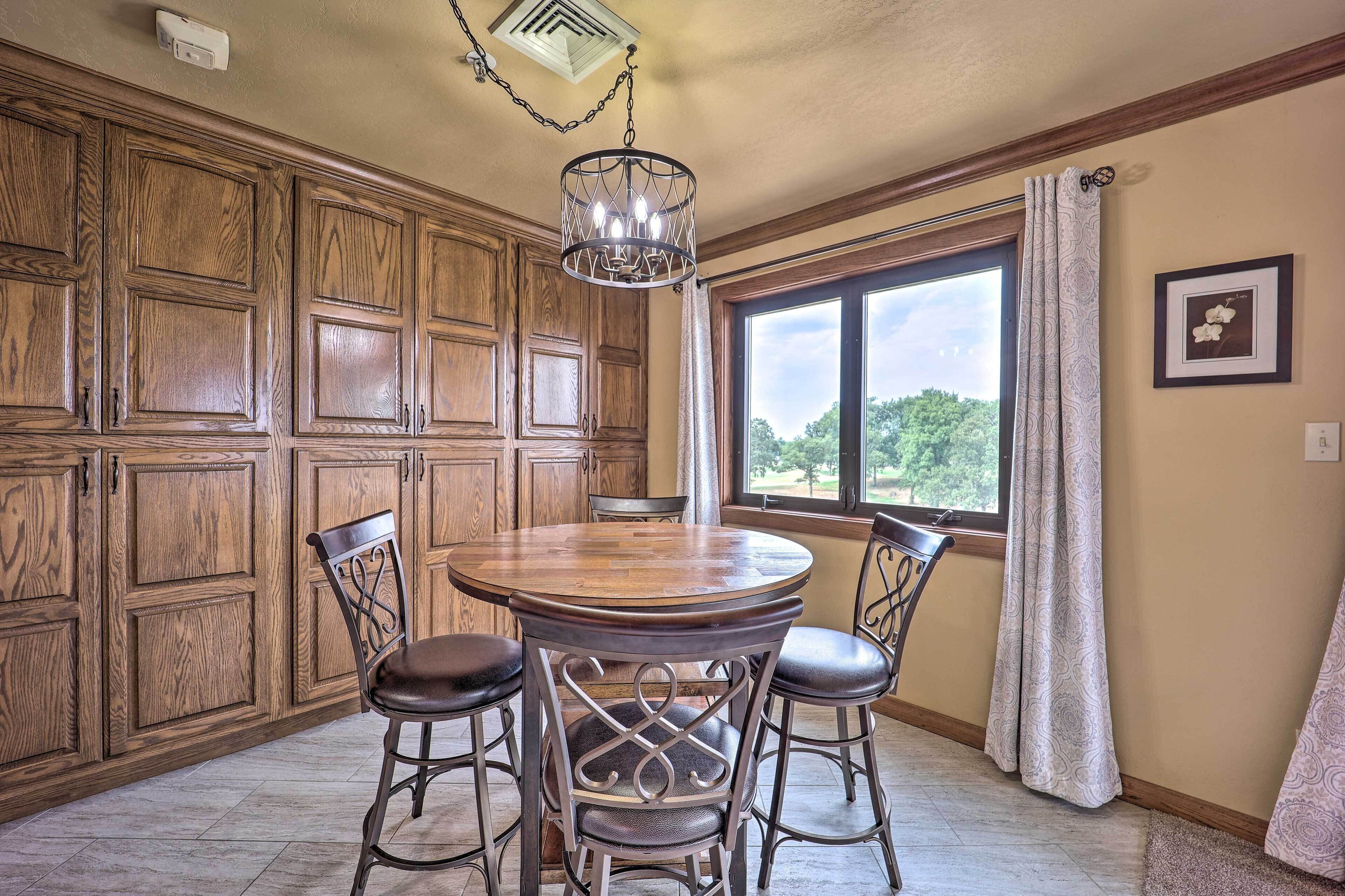 A dining area with a round wooden table and four chairs, wooden paneling on the walls, a window with a view, and a hanging light fixture.